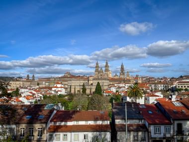 Panoramablick auf Santiago de Compostela mit den Türmen der Kathedrale über den roten Ziegeldächern unter blauem Himmel mit weißen Wolken.