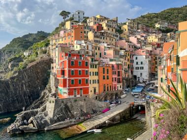 Riomaggiore village with colorful houses stacked on cliffs above the sea. Boats rest in a small harbor, surrounded by green hills under blue sky.
