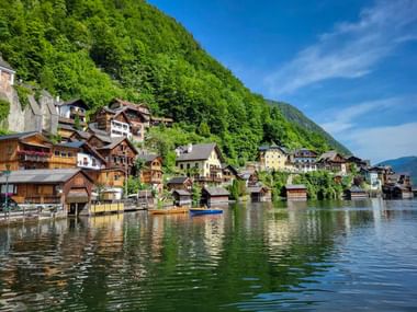 Traditional alpine houses of Hallstatt reflected in calm lake waters, with forested mountains rising steeply behind the village under blue sky.