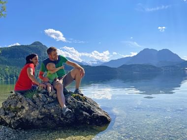 Family of three sitting on a rock by crystal-clear Altausseer See in Salzkammergut, with Dachstein mountains reflected in calm water.