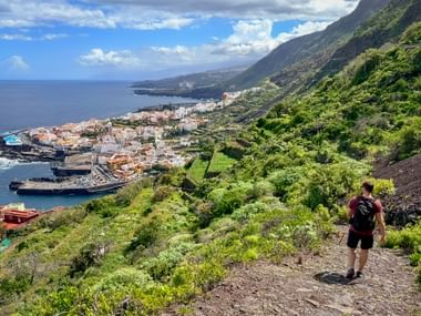 Wanderer mit Rucksack auf Bergpfad mit Blick auf die Küstenstadt Garachico auf Teneriffa, mit grünem Hang, Meer und blauem Himmel.