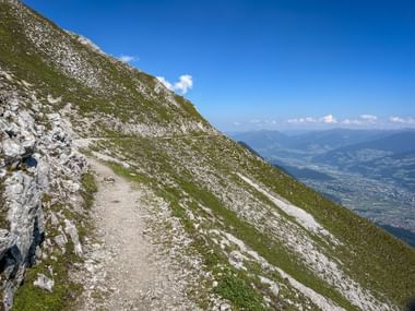 Schotterwanderweg an steilem Berghang nahe der Hafelekarspitze mit felsigem Gelände, grünem Gras und Panoramablick auf das Inntal.