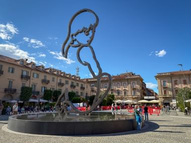 Large metal sculpture of a stylized figure in a circular fountain basin on a town square in Alba, Piedmont, with historic buildings in background.