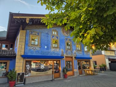 Blaues historisches Gebäude mit kunstvollen Fassadenmalereien in Garmisch. Das zweistöckige Haus zeigt dekorative Fresken und moderne Geschäfte im Erdgeschoss.