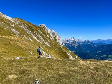 Hiker with backpack on grassy alpine slope in Tennengebirge with rocky peaks and mountain ranges under blue sky.