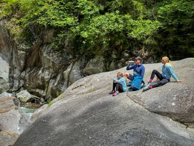 Family of three hikers resting on smooth rock beside mountain stream in Passer Gorge. Green vegetation covers rocky cliffs above the water.