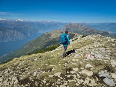 Wanderer in blauer Jacke auf dem Monte Baldo Grat mit Blick auf den Gardasee und schneebedeckte Alpen unter blauem Himmel.