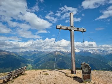 Hölzernes Gipfelkreuz am Blaser im Gschnitztal mit Bergpanorama. Schneebedeckte Gipfel in der Ferne unter blauem Himmel mit Wolken.