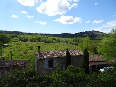Steinbauernhaus mit Terrakottadach umgeben von grünen Weinbergen in der Provence. Sanfte Hügel und ein Bergdorf in der Ferne sichtbar.