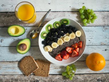 Bowl of yoghurt with kiwis, blackberries, bananas, strawberries, chia seeds and wholemeal bread with a sliced avocado, a glass of orange juice