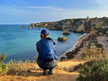 Hiker with blue backpack overlooking Praia da Dona Ana beach in Algarve, with golden cliffs, rock formations, and turquoise water.