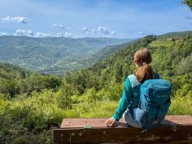 Woman with blue backpack sitting on wooden bench overlooking green valley in Piedmont. Forested hills and mountains under blue sky.