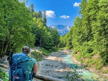 Wanderer mit Rucksack blickt auf türkisfarbenen Bach in der Partnachklamm, umgeben von grünem Wald mit Zugspitze im Hintergrund.