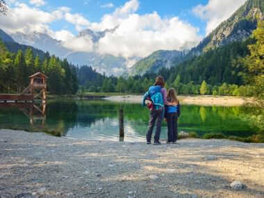 Zwei Wanderer am Ufer des Jasna-Sees mit Holzbootshaus, umgeben von bewaldeten Bergen und dramatischen Wolken in den Julischen Alpen.