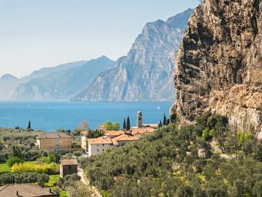 Torbole mit Kirchturm zwischen dramatischen Felsen und dem Gardasee, umgeben von Olivenhainen und mediterraner Vegetation.