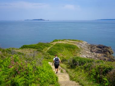 Wanderer mit Rucksack auf Küstenpfad umgeben von grüner Vegetation und rosa Blüten, mit Blick auf blaues Meer und entfernte Inseln.