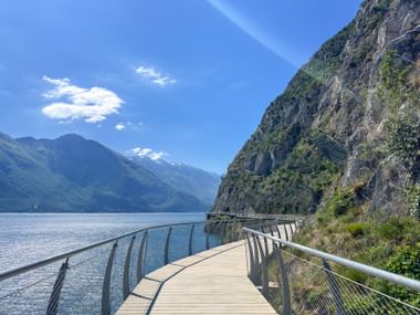 Holzsteg am Gardasee mit Metallgeländer, steile Felswand rechts, Berge über blauem Wasser unter klarem Himmel.