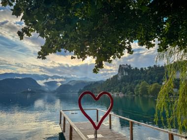 Rote Herzskulptur auf Holzsteg am Bleder See. Die Burg Bled thront auf einer bewaldeten Klippe über dem ruhigen türkisfarbenen Wasser.
