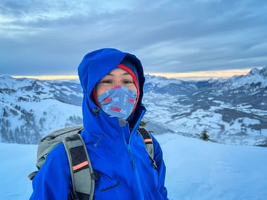 Person in blauer Winterjacke und Gesichtsmaske mit Rucksack auf verschneitem Gipfel in Salzburg, mit schneebedeckten Gipfeln und Tal.