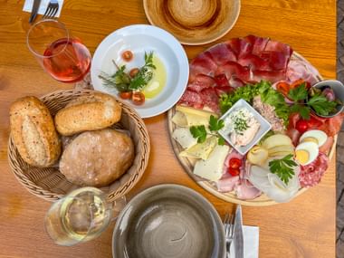 Wooden table with South Tyrolean snack platter featuring cold cuts, cheese, eggs, bread basket, wine glasses, and decorative straw hat.