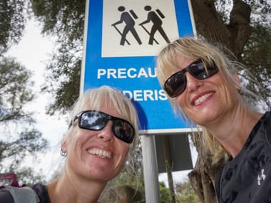 Two smiling blonde women with sunglasses pose in front of a blue hiking trail sign showing hiker symbols, surrounded by trees.