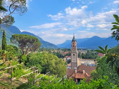 View from Tappeinerweg in Meran showing a church tower amid lush vegetation, with mountains and blue sky in the background.