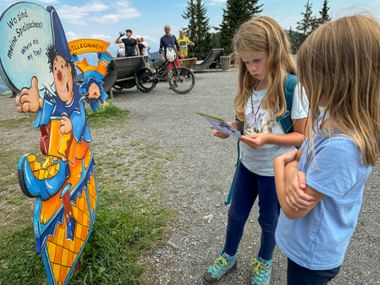 Two girls reading a map at Montelino's adventure trail starting station in Saalbach-Hinterglemm, with a colorful cartoon cutout and families in background.