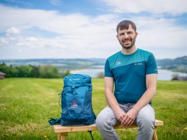 Man in turquoise Eurohike shirt sitting on wooden bench with blue Vaude backpack, green meadow and lake landscape in background.