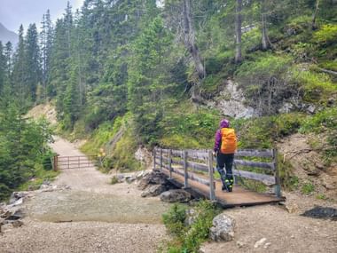 Wanderin in lila Jacke mit orangem Rucksack überquert Holzbrücke auf Bergweg bei der Zugspitze, umgeben von grünem Wald und Felsen.