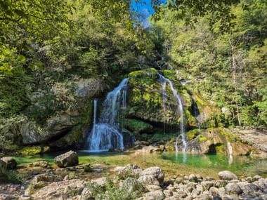 Virje-Wasserfall im Soča-Tal fließt über moosbedeckte Felsen in ein klares türkisfarbenes Becken, umgeben von üppigem Wald und Felsgelände.