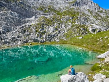 Wanderer mit Rucksack sitzt auf Felsen neben kristallklarem türkisen Bergsee, umgeben von dramatischen Kalksteinfelsen und grünen Wiesen.