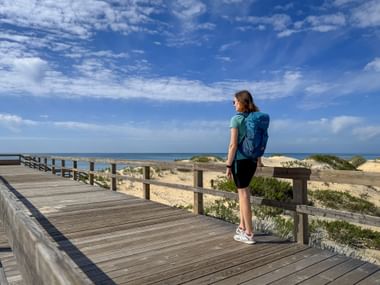 Woman with backpack standing on wooden boardwalk overlooking sandy dunes and ocean at Praia Verdelago, Algarve under blue sky with clouds.