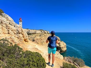 Wanderer mit Rucksack auf Klippenpfad an der Algarve, Portugal. Weißer Leuchtturm auf felsigem Kap, türkisfarbenes Meer und blauer Himmel.