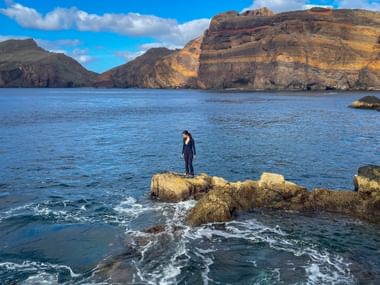 Woman standing on coastal rocks at São Lourenço peninsula, Madeira, with dramatic layered cliffs and blue ocean under sunny skies.