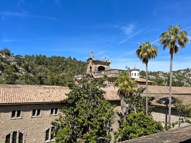 Stone buildings of Lluc Monastery with terracotta roofs, palm trees, and mountains under blue sky in Mallorca on the GR221 hiking trail.