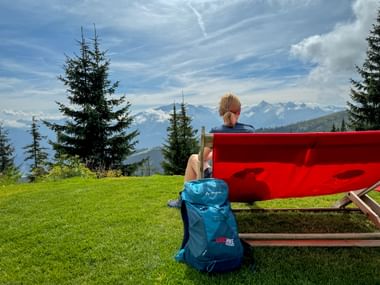 Person on red bench overlooking alpine meadow with blue backpack, pine trees, and snow-capped mountains in Pinzgau region.