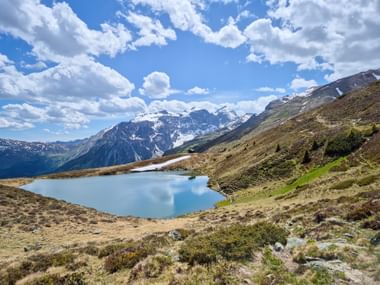Türkisfarbener Bergsee umgeben von Almwiesen mit schneebedeckten Gipfeln im Hintergrund unter blauem Himmel mit weißen Wolken im Gschnitztal-Obernberg.