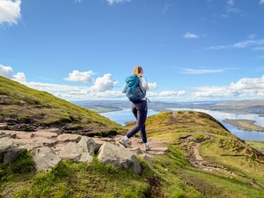 Wanderin mit Rucksack auf felsigem Pfad auf grünem Hügel mit Blick auf Loch Lomond unter blauem Himmel mit weißen Wolken.