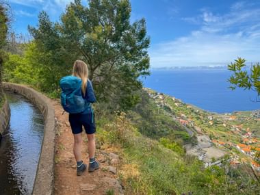 Wanderin mit blauem Rucksack auf einem Levada-Weg auf Madeira mit Blick auf das Küstendorf Câmara de Lobos und den Atlantik.