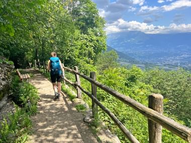 Hiker with blue backpack walking on mountain trail with wooden fence, overlooking Meran valley and mountains in South Tyrol under blue sky.