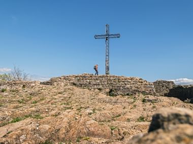 Hiker standing near tall metal summit cross on rocky hilltop at Rocca di Manerba. Ancient stone walls visible on rocky terrain under blue sky.