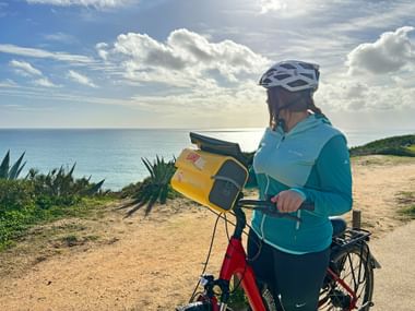 Radfahrer in türkisfarbener Jacke und Helm liest eine Karte auf einem Küstenweg mit Blick auf den Atlantik an der Algarve, Portugal.