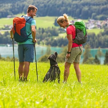 Two hikers with backpacks and a black dog on a green meadow in Salzkammergut, overlooking a turquoise lake and mountain village.