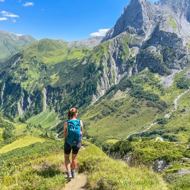 Hiker with blue backpack walking on mountain trail overlooking green valleys and rocky peaks in the Allgäu-Lechtal Alps on the Arlberg Trail.