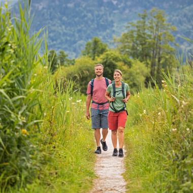 Zwei Wanderer auf einem Schotterweg durch hohes Gras nahe dem Wolfgangsee im Salzkammergut, mit bewaldeten Bergen im Hintergrund.