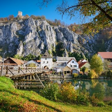 Dorf Essing mit Holzbrücke über Fluss, traditionellen Häusern und dramatischen weißen Kalksteinfelsen im Altmühltal unter blauem Himmel.