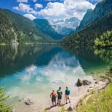 Three hikers with backpacks standing at the shore of Gosausee lake in Salzkammergut, with the snow-capped Dachstein mountains reflected in the water.