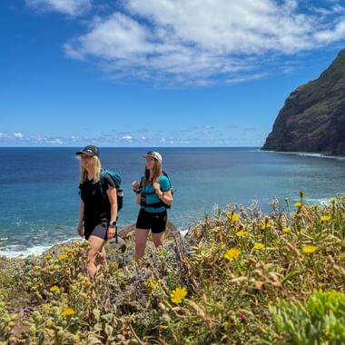 Two female hikers with backpacks standing on a coastal cliff in Madeira, surrounded by yellow wildflowers, overlooking the blue ocean.