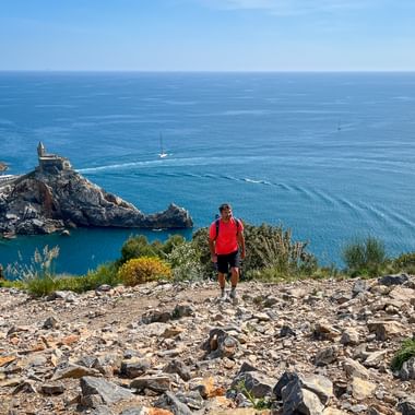 Hiker in red jacket on rocky trail overlooking Porto Venere bay with church on promontory, blue Mediterranean Sea, and sailboats.
