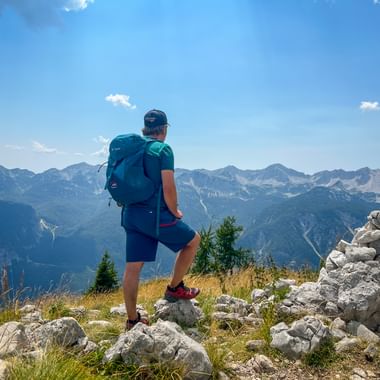 Hiker with backpack standing on rocky summit in the Julian Alps, overlooking mountain ranges under blue sky with stone cairn nearby.